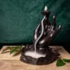 Тамянник (кадилница) с обърнат поток Lotus hand, оформен като ръка, с дим, който се стича по пръстите му, разположен върху дървена повърхност със зелен лист на заден план.