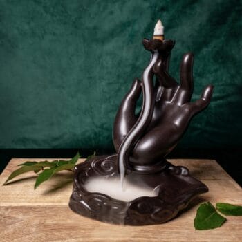 Тамянник (кадилница) с обърнат поток Lotus hand, оформен като ръка, с дим, който се стича по пръстите му, разположен върху дървена повърхност със зелен лист на заден план.