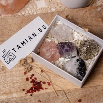 Кутия с етикет "Подаръчна кутия с 5 минерални камъни" съдържа пет необработени кристала, разположени върху нарязана хартия. Придружаваща кутията картичка с надпис "TAMIAN.BG" лежи върху дървена повърхност, украсена със сухи цветя и червени кристали.