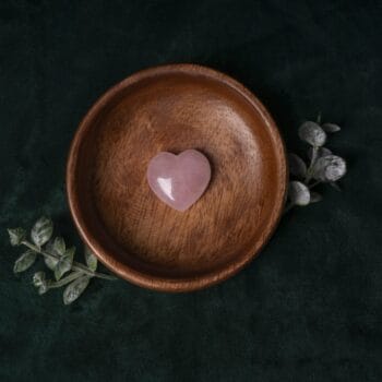 Дървена купа с Камък Розов кварц - сърце е поставена върху тъмнозелен плат с листа в близост.