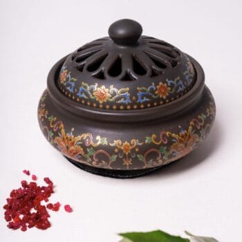 Бутиков тамянник Екзотика 3v1, включващ сложни шарки, лежи върху бяла повърхност с червени парчета смола, поставени до нея.