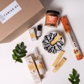 Плоско оформление на Комплект Пало санто, включващо дървени пръчици Palo Santo, чиния, спрей бутилка, прах, етерично масло и ароматни пръчици, изложени до брандирана кутия на Tamian.bg и зелени листа.