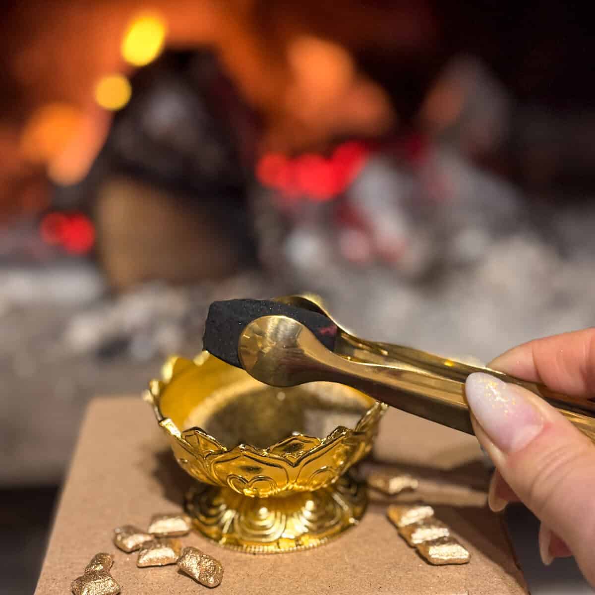 Ръката държи клещи с дървени въглища над Комплект Златна Вечер златна кадилница, а въглените нежно светят на размазания фон.