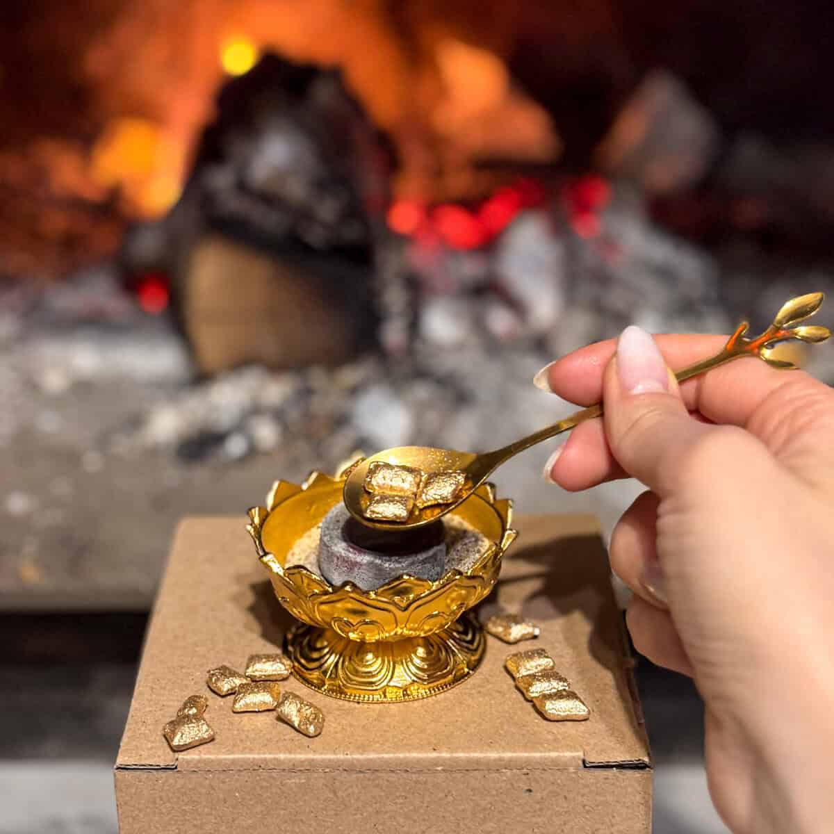 Ръка държи златна лъжица със златни парчета над декоративната Комплект Златна Вечер кадилница, на фона на горящ огън и дървени трупи.